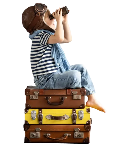 This image features a young child dressed in a playful, adventure-themed outfit, sitting atop a stack of vintage suitcases. The child is wearing a striped shirt and denim overalls, with a pilot's cap and goggles on their head, and is holding a camera up to their face as if taking a picture. The cap and goggles suggest an old-fashioned aviator theme. The child’s bare feet dangle off the side of the suitcases, adding to the relaxed and imaginative scene. The background is transparent, which emphasizes the child and the suitcases, evoking a sense of travel and exploration.