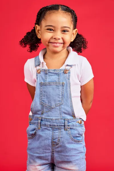 The image shows a young girl with a joyful expression, standing against a vibrant red background. She has curly black hair pulled back into two puff ponytails and is smiling broadly. The girl is wearing a white short-sleeved shirt and blue denim overalls. Her hands are casually placed in the pockets of her overalls, and the straps are secured with classic overall buckles. Her cheerful demeanor and casual pose give the picture a friendly and approachable feel.